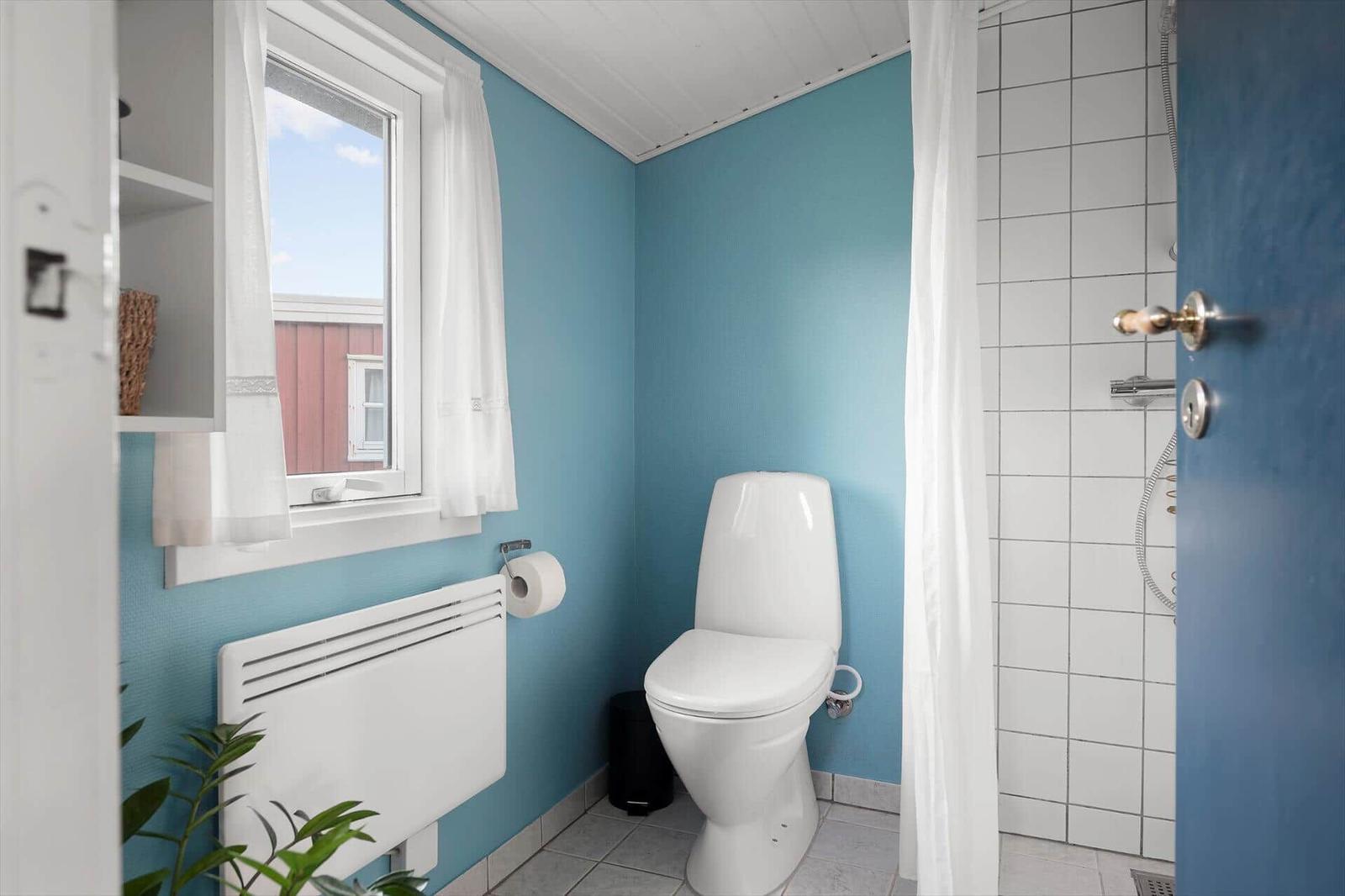 Badezimmer mit Toilette, Dusche und Fenster mit weißen Vorhängen.