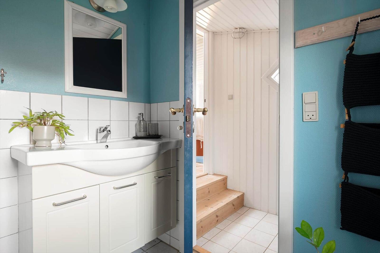 Badezimmer mit Waschbecken, Spiegel und Pflanze. Tür führt in einen Raum mit Holzstufen.