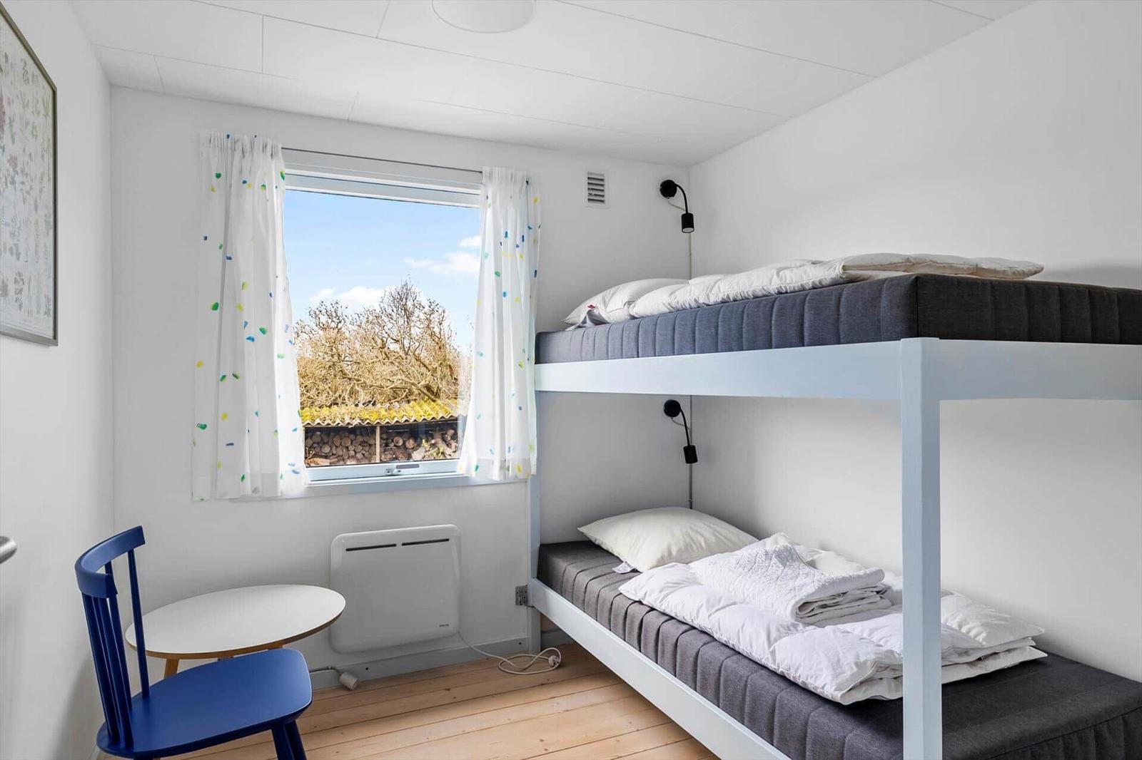 Zimmer mit Doppelstockbett, Fenster mit Vorhängen und kleinem Tisch mit Stuhl.