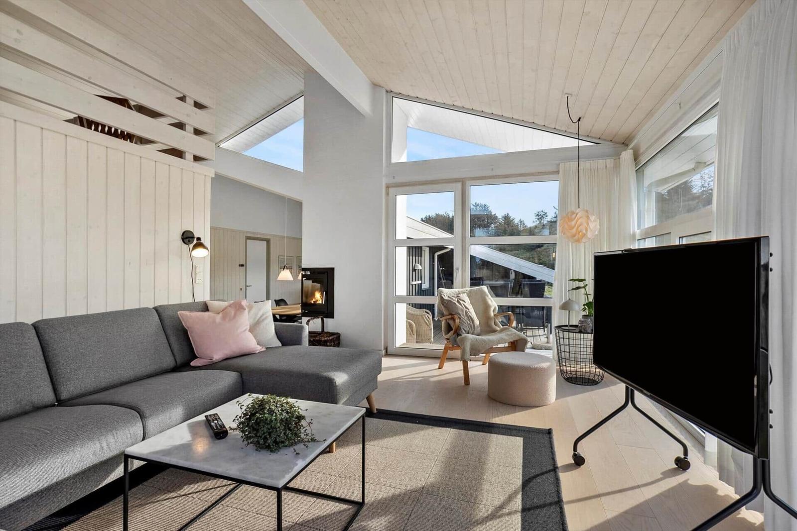 Wohnzimmer mit Sofa, Fernseher und großem Fenster. Holzdecke und Kamin sichtbar.
