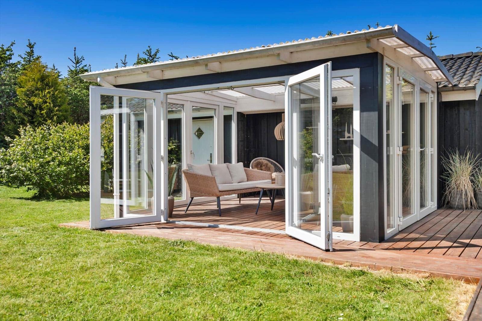 Terrasse mit Glaswänden und Sitzmöbeln auf Holzdecke. Grasfläche und Bäume im Hintergrund.
