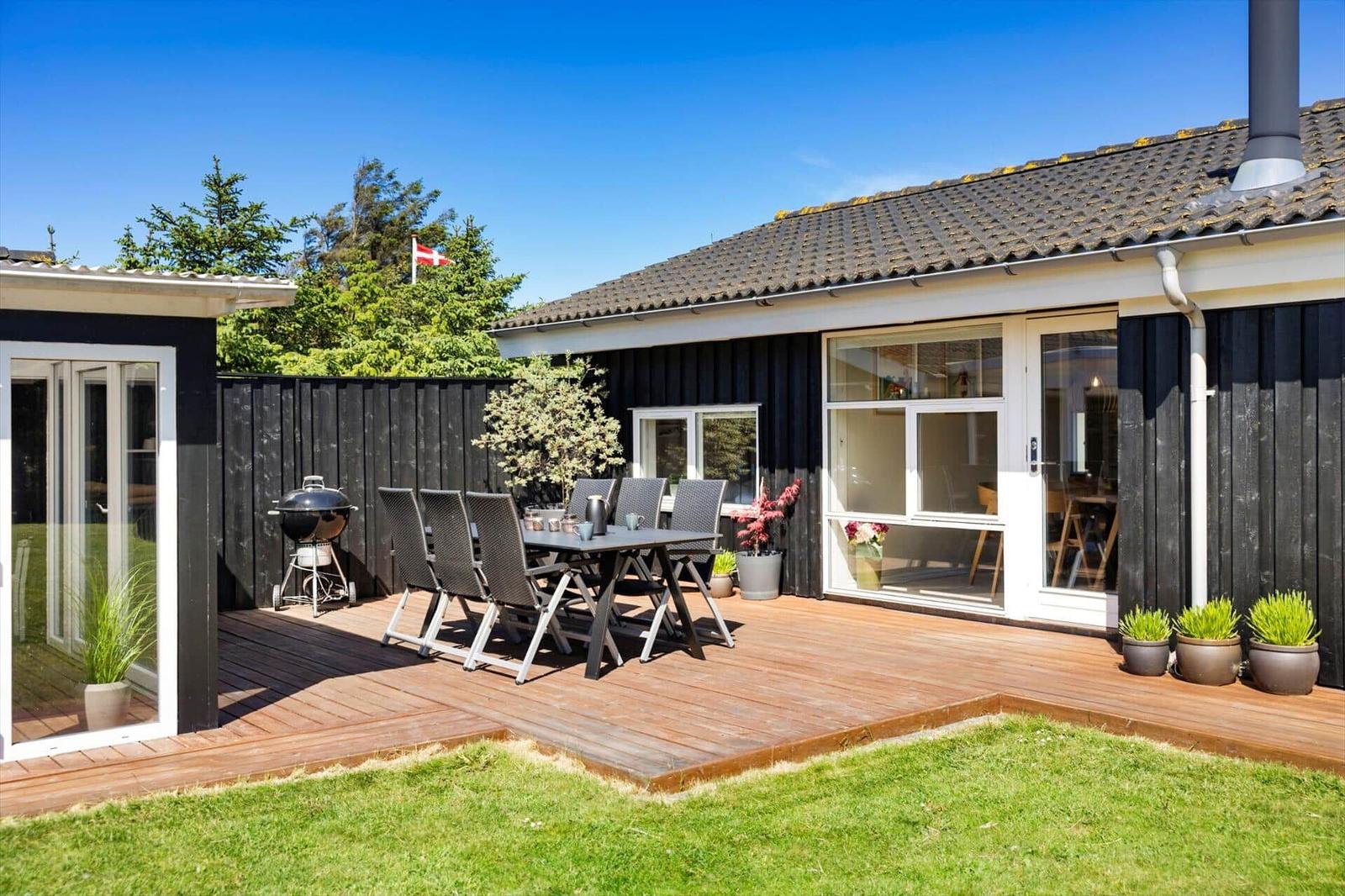 Terrasse mit Tisch, Stühlen und Grill. Haus mit Fenstern und Pflanzen.
