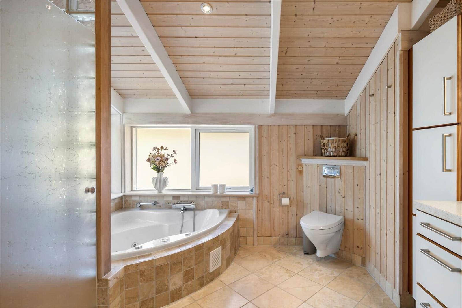 Bad mit Whirlpool, Toilette und Holzwandverkleidung. Fenster mit Blumenvase.