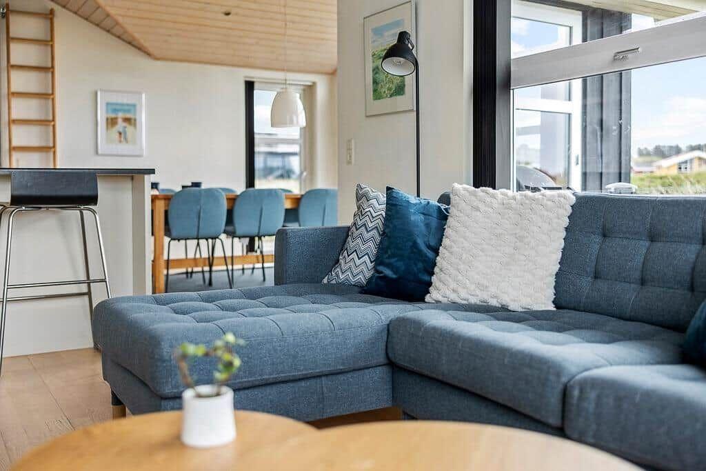 Ein Wohnzimmer mit blauem Sofa, Kissen und Holztisch. Im Hintergrund ist eine Küche mit Stühlen.