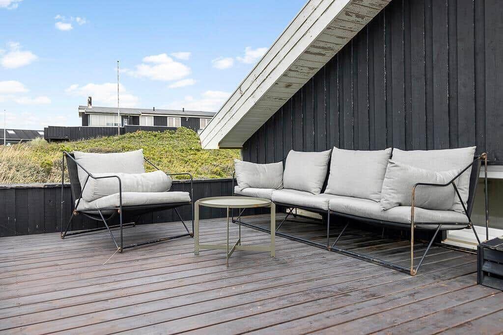 Terrasse mit Sofa und Sessel, Holzboden, schwarze Wand, Blick auf Grünfläche.