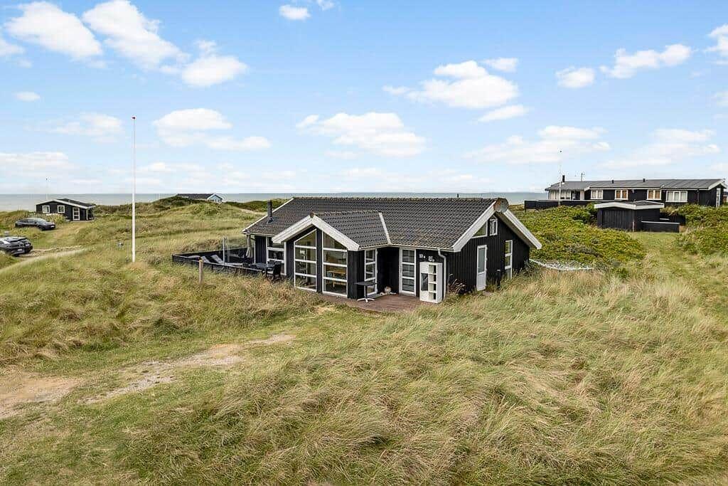 Schwarzes Strandhaus mit großem Fenster und Terrasse zwischen Dünen und Meer.
