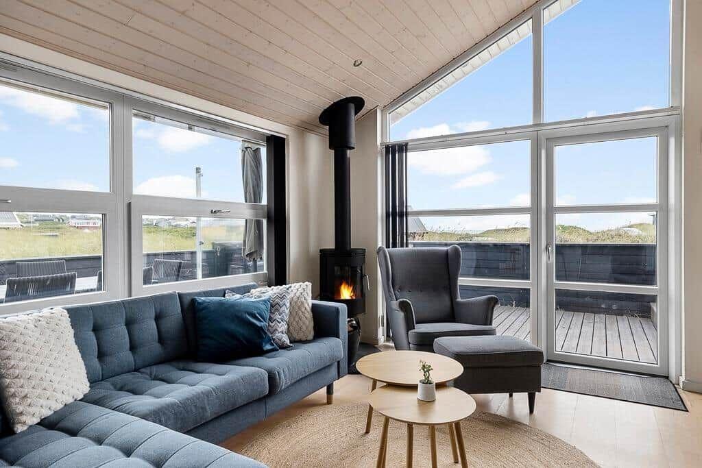 Ein Wohnzimmer mit Sofa, Sessel und Kamin. Große Fenster bieten Blick auf die Terrasse.