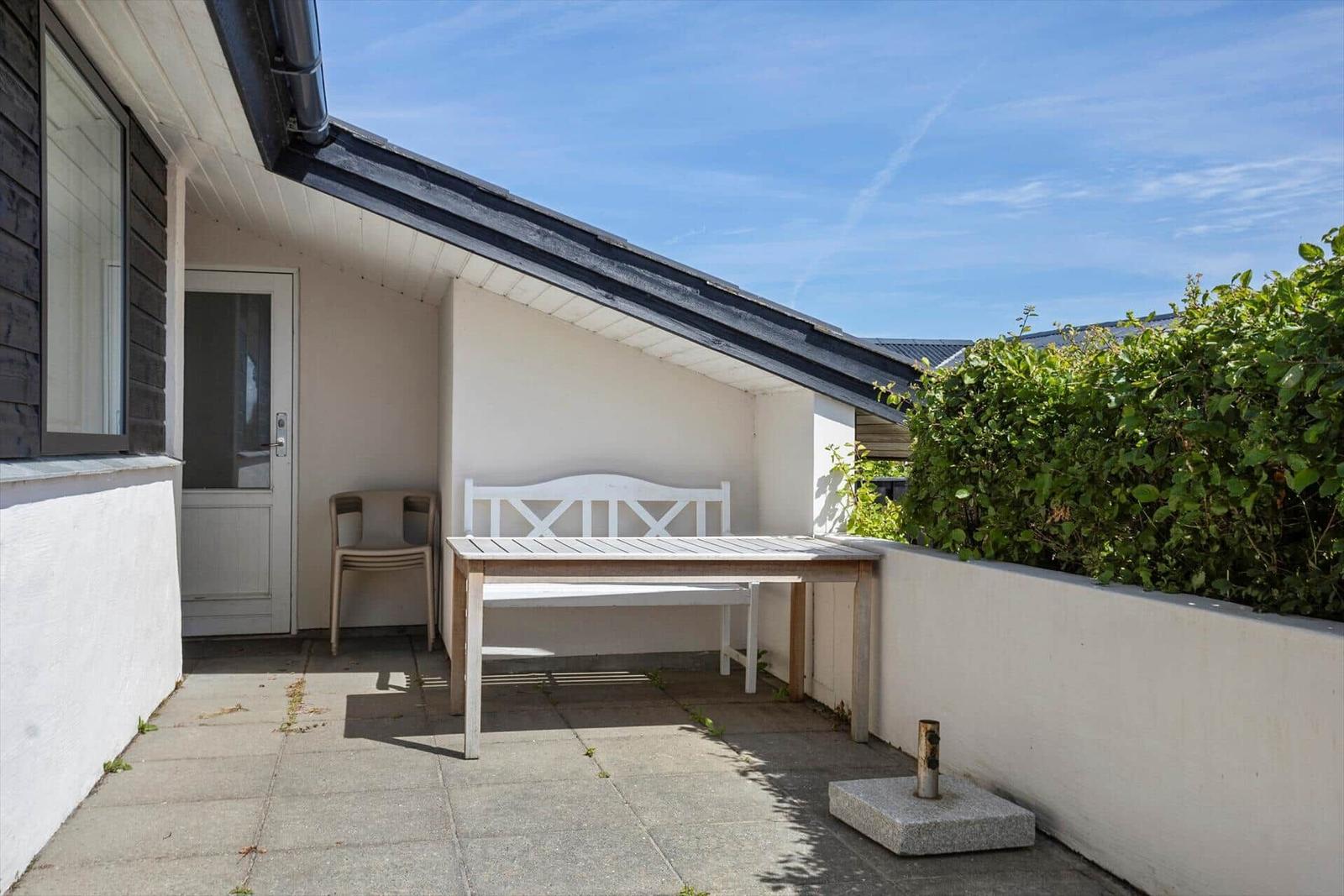Terrasse mit Tisch, Bank und Stuhl unter einem Dach. Hintergrund: grüne Hecke und blauer Himmel.