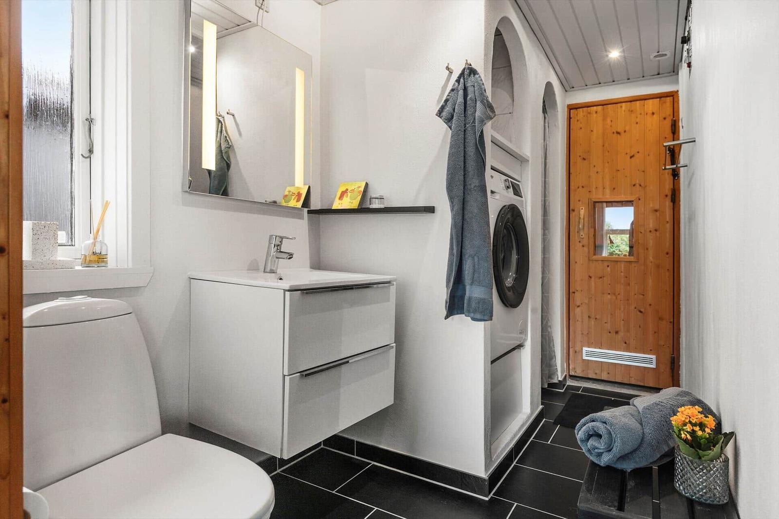 Badezimmer mit Waschmaschine, Spiegelschrank und Toilette. Schwarze Fliesen und Holztür.