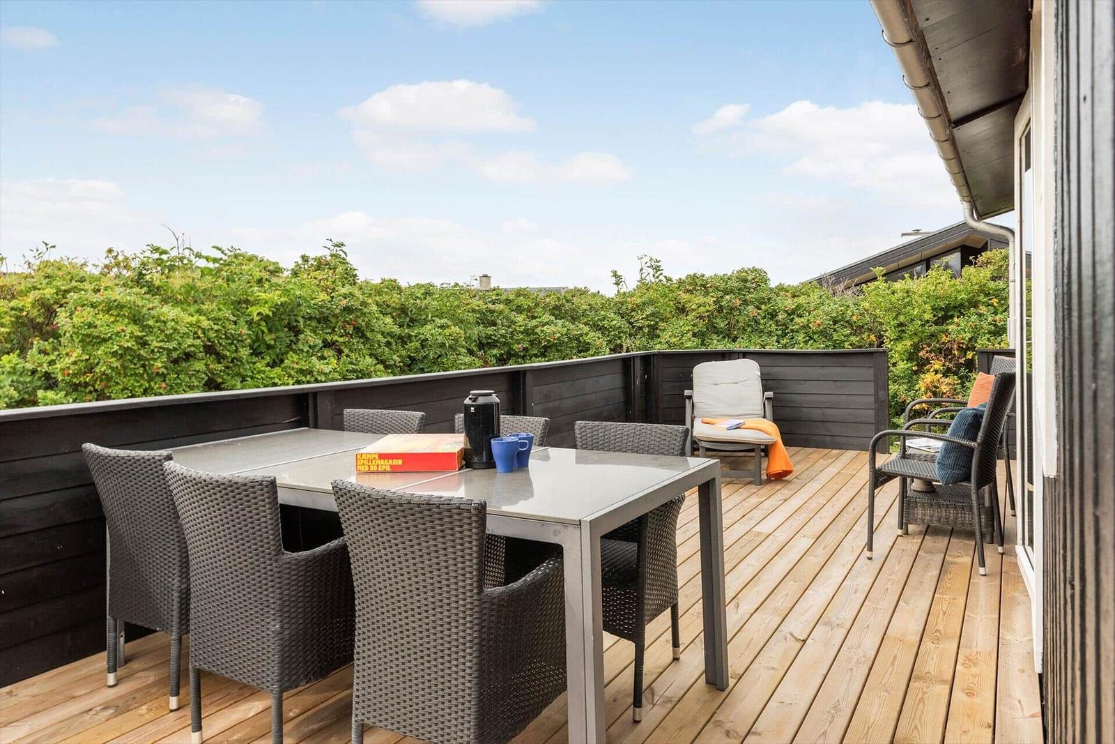 Terrasse mit Esstisch, Stühlen und Sitzgelegenheit. Blick auf grüne Hecke und Himmel.