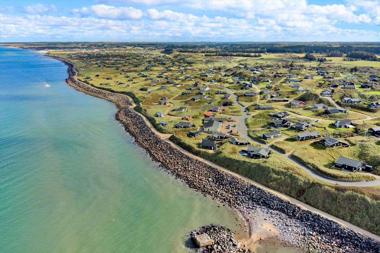 Aerial view of coastal village with houses on grassy dunes beside calm sea.