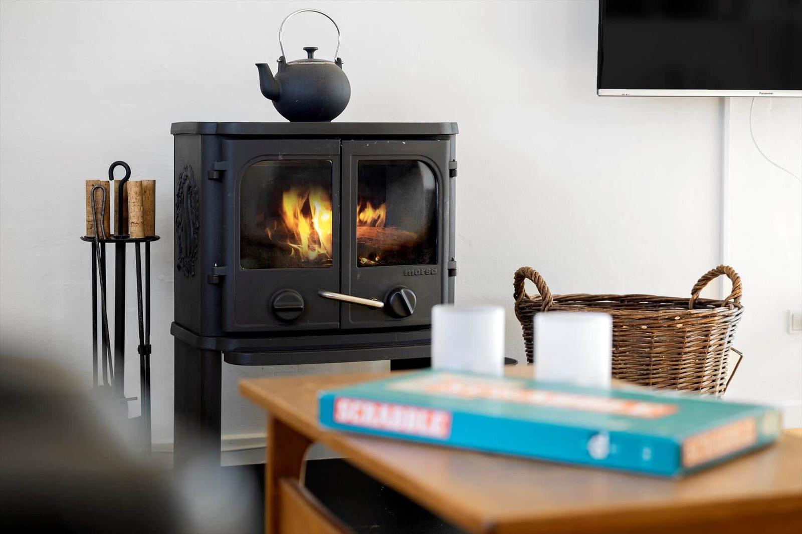 Ein Kaminofen mit brennendem Holz, darauf ein Teekessel, daneben ein Korb und eine Fernsehleinwand.