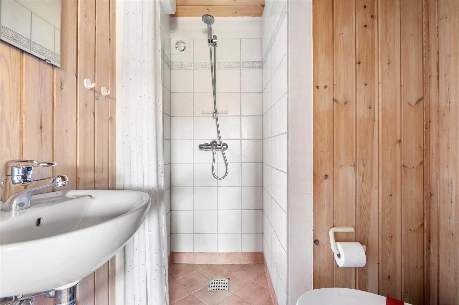 Badezimmer mit Dusche, Waschbecken und Toilette. Holzwände und Fliesenboden.