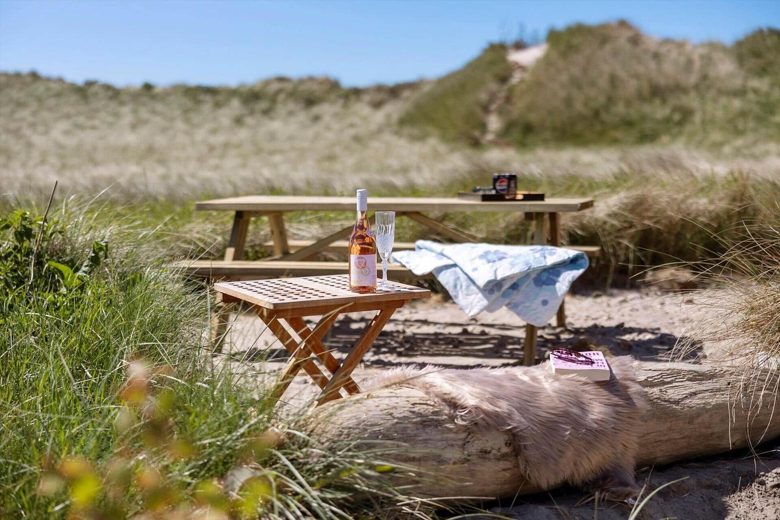Holztisch mit Weinflasche und Glas auf Strand. Hintergrund: Dünen und Gras.