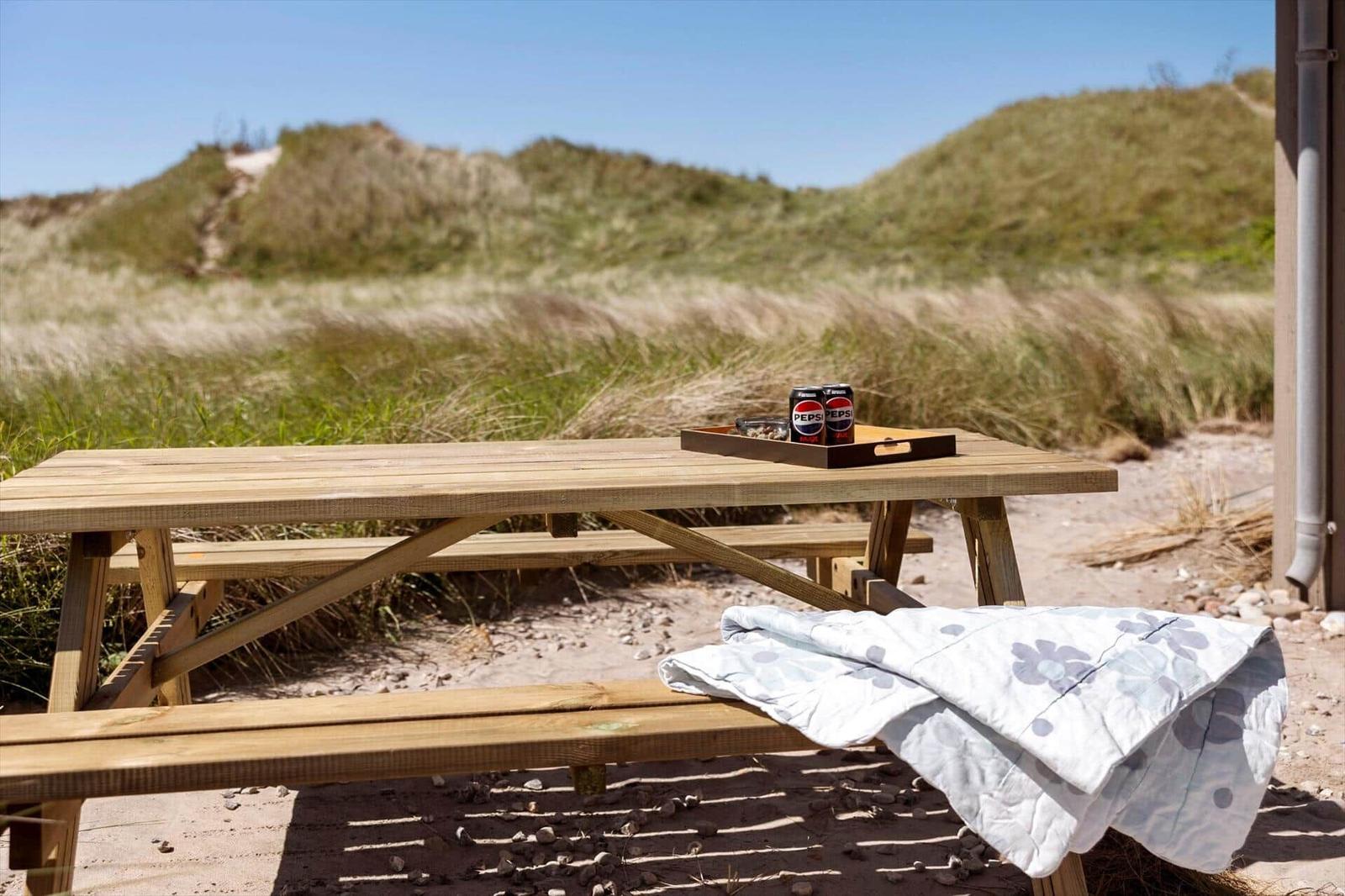 Holzpicknicktisch mit Decke und Getränken vor Dünen.
