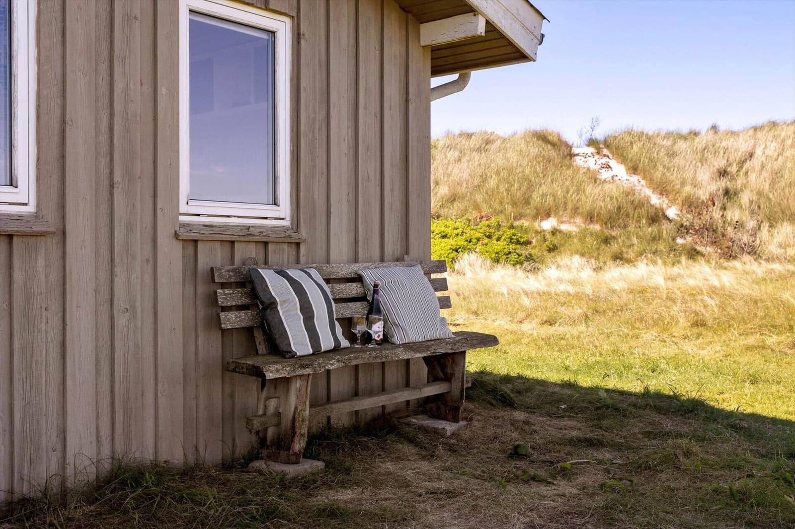 Holzbank mit Kissen vor einem Haus mit Dachüberstand und Blick auf Dünen.