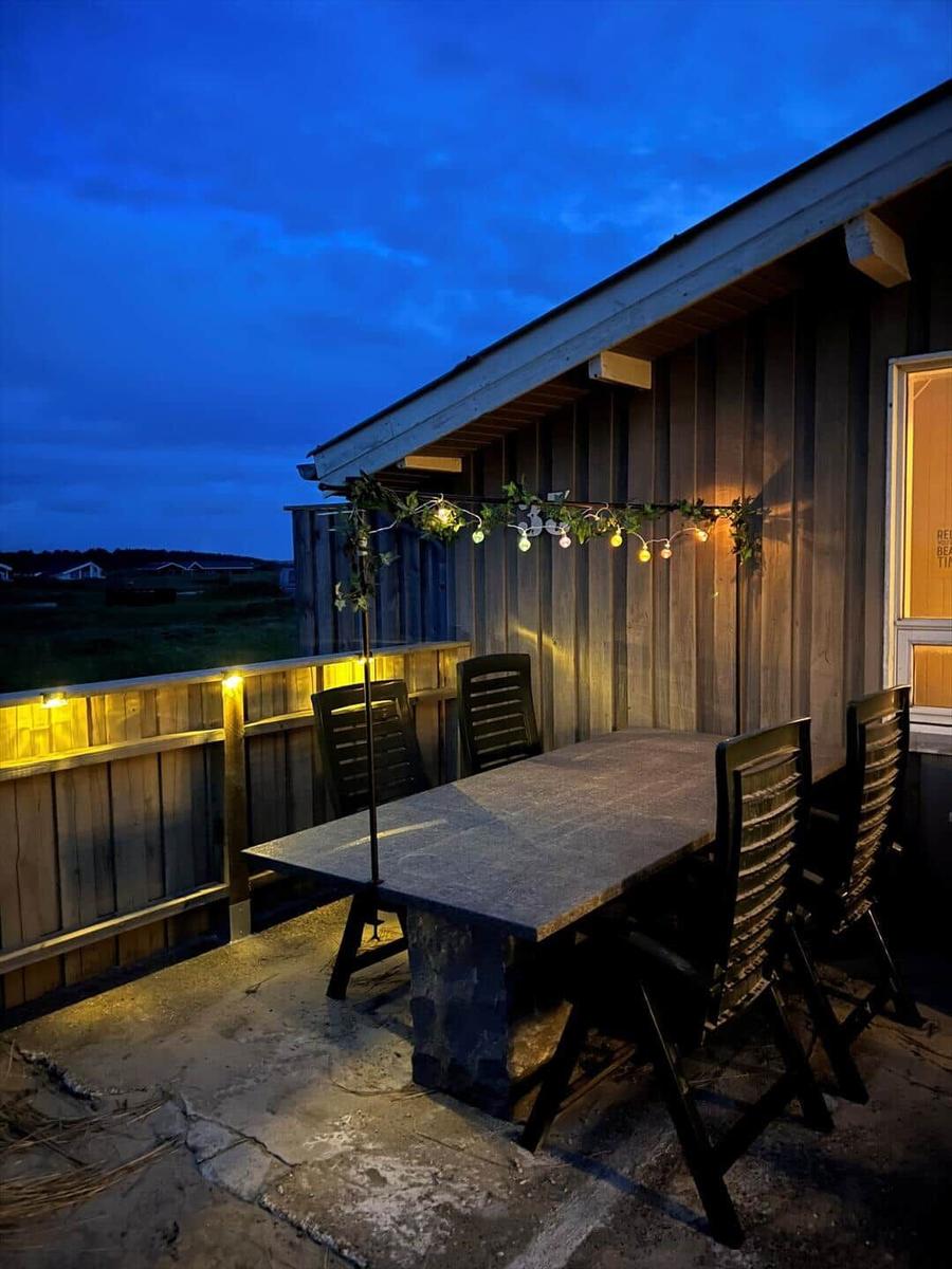 Terrasse mit Tisch und Stühlen, beleuchtet von Girlanden und Wandlampen bei Dämmerung.