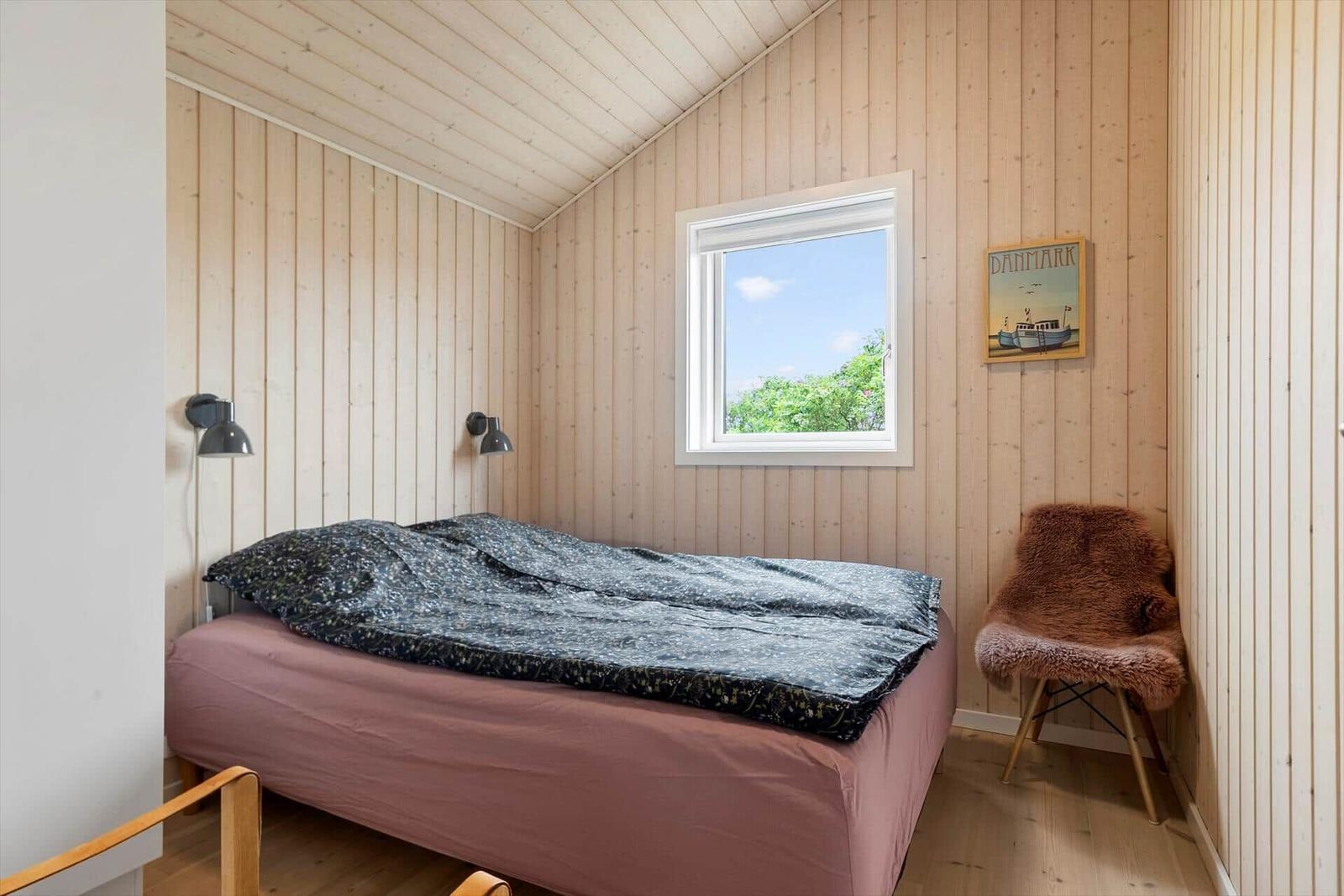 Schlafzimmer mit Holzwand, Bett, Fenster und Stuhl mit Fellbezug.