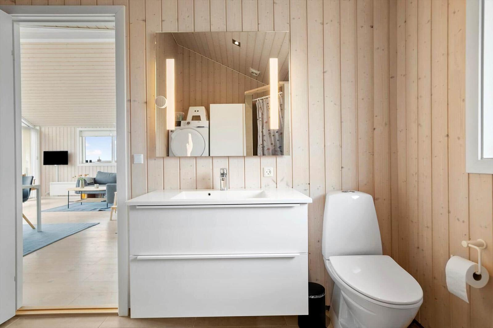 Badezimmer mit Waschbecken, Toilette und Spiegel. Holzwand und Blick in Wohnzimmer.