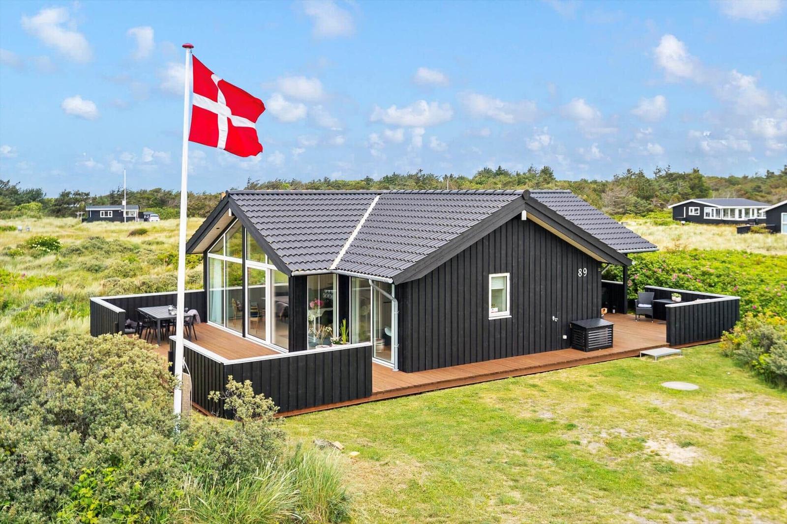 Schwarzes Ferienhaus mit Terrasse und dänischer Flagge auf grünem Grund.