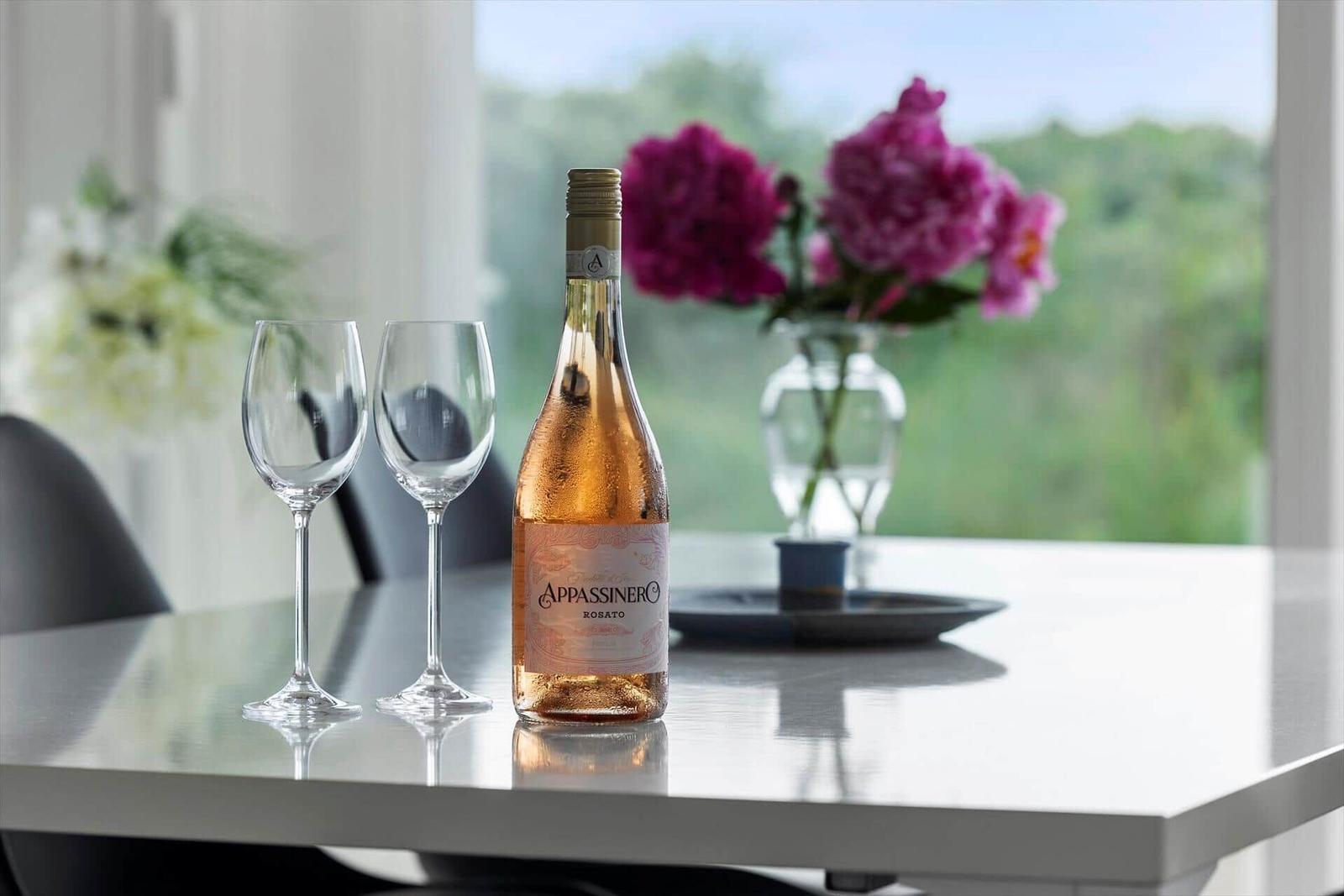 Auf dem Tisch steht eine Flasche Roséwein mit zwei Gläsern. Im Hintergrund Blumen und ein Fenster.
