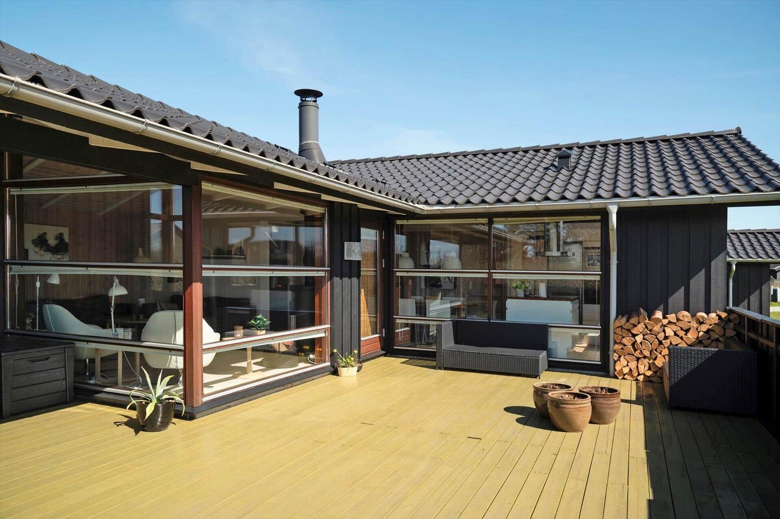 Terrasse mit Holzdecke, Sitzgelegenheiten und Holzstapel. Glaswände zeigen Innenräume.