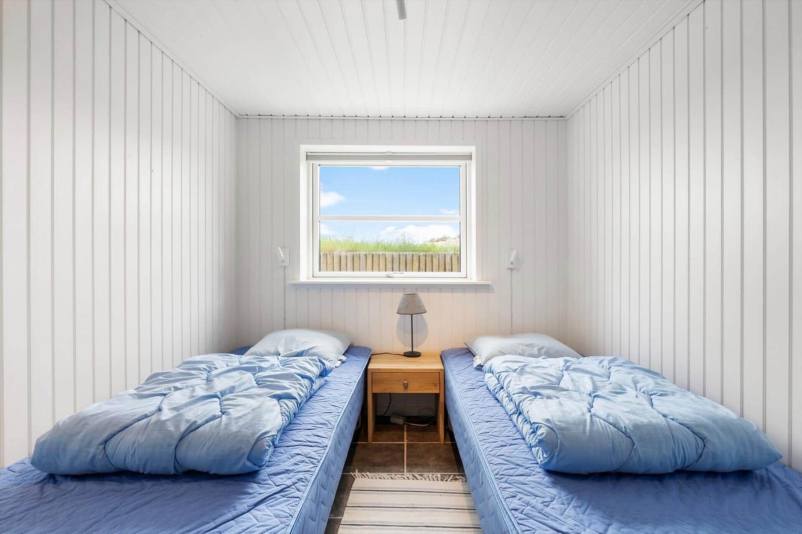 Two beds with blue bedding are in a room with white walls and a window.