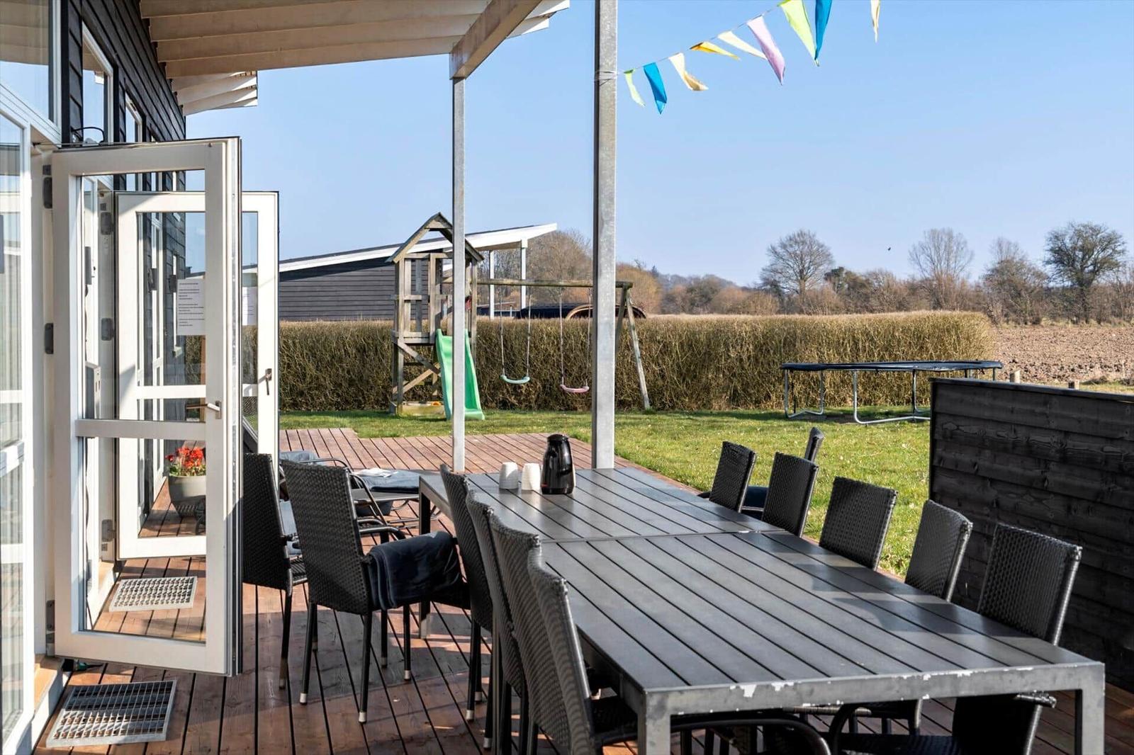 Terrace with table and chairs, playground and trampoline in the background.