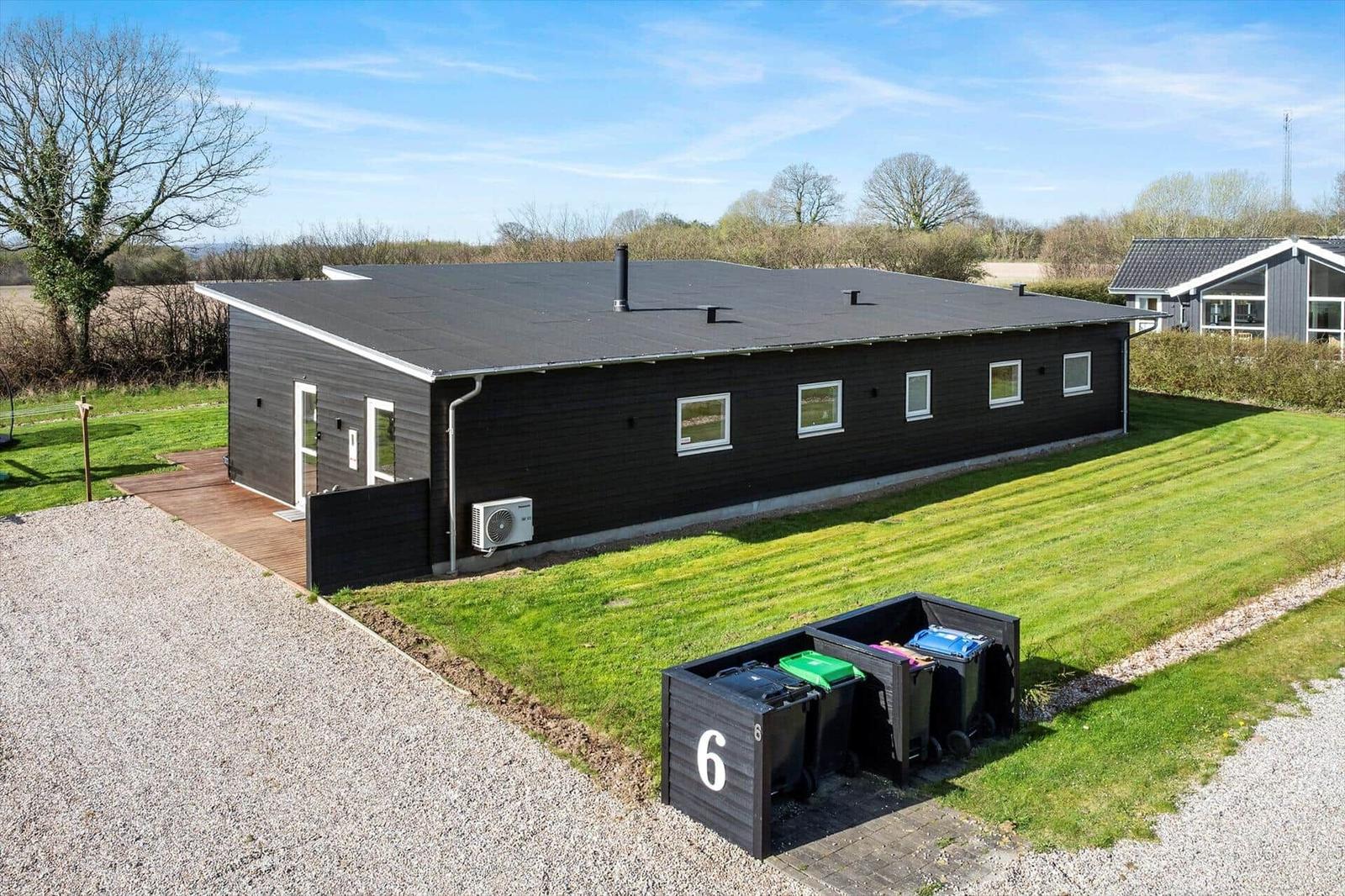 Black vacation house with green lawn and trash bins. Entrance with wooden terrace.