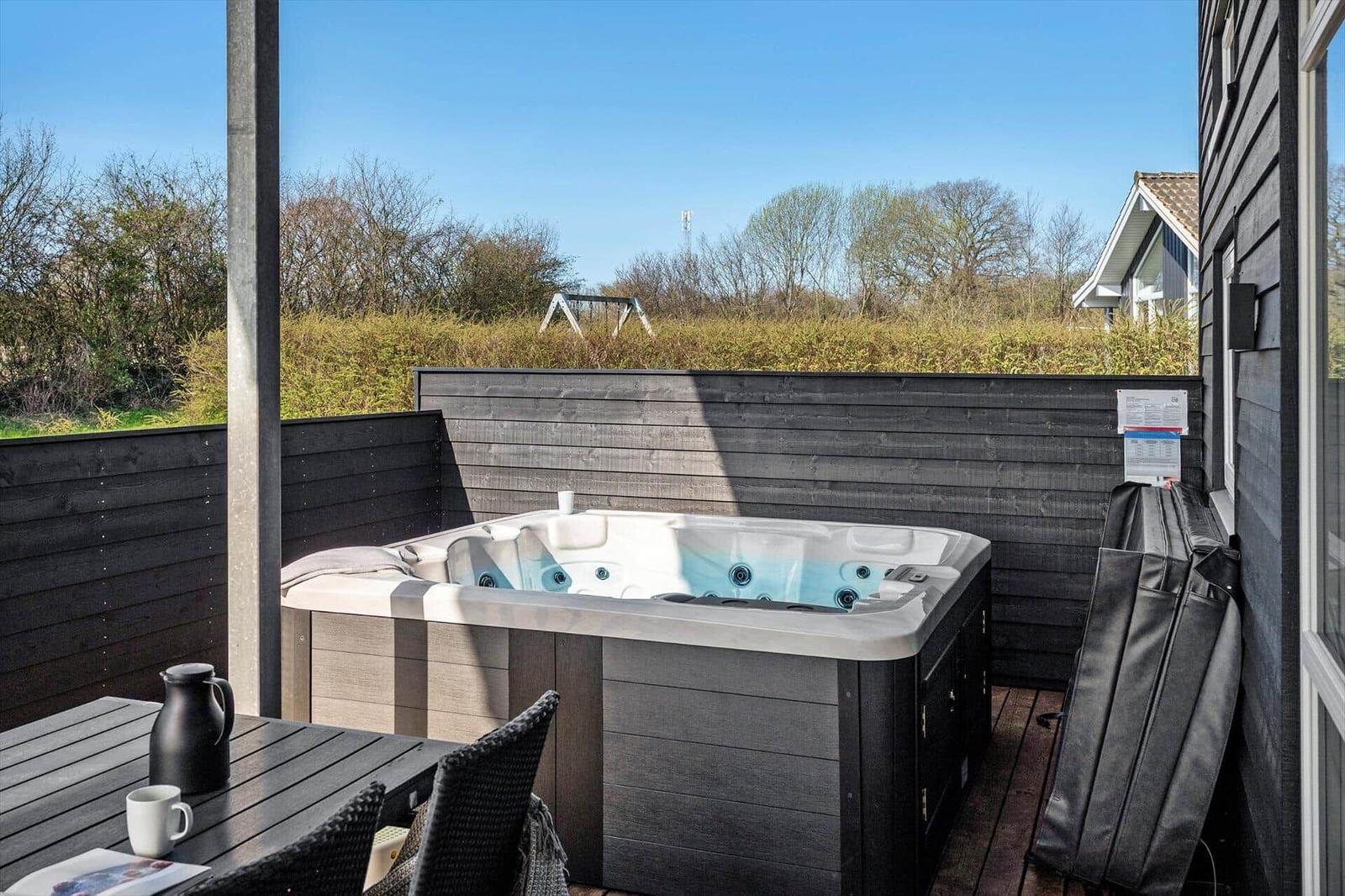 Outdoor hot tub with table and chairs on terrace. Background: green area and wooden fence.