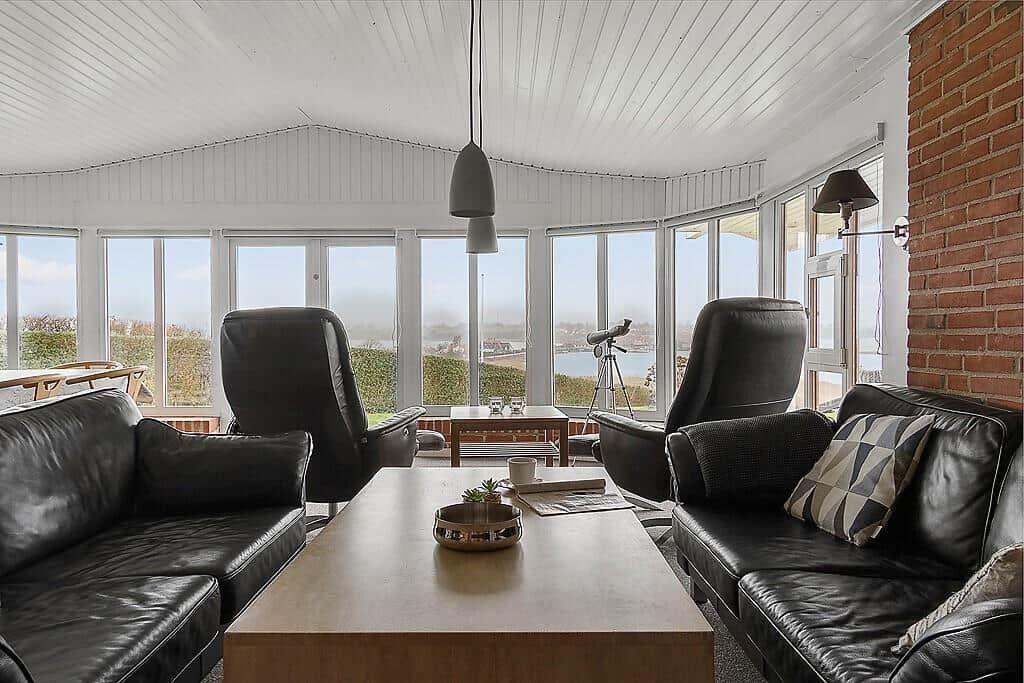 Wohnzimmer mit schwarzen Ledermöbeln, Holztisch und Blick auf Wasser durch große Fenster.