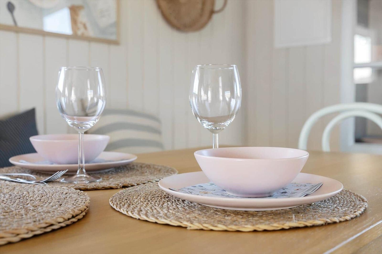 Table with two plates, bowls, glasses, and placemats. Background with white walls and chairs.
