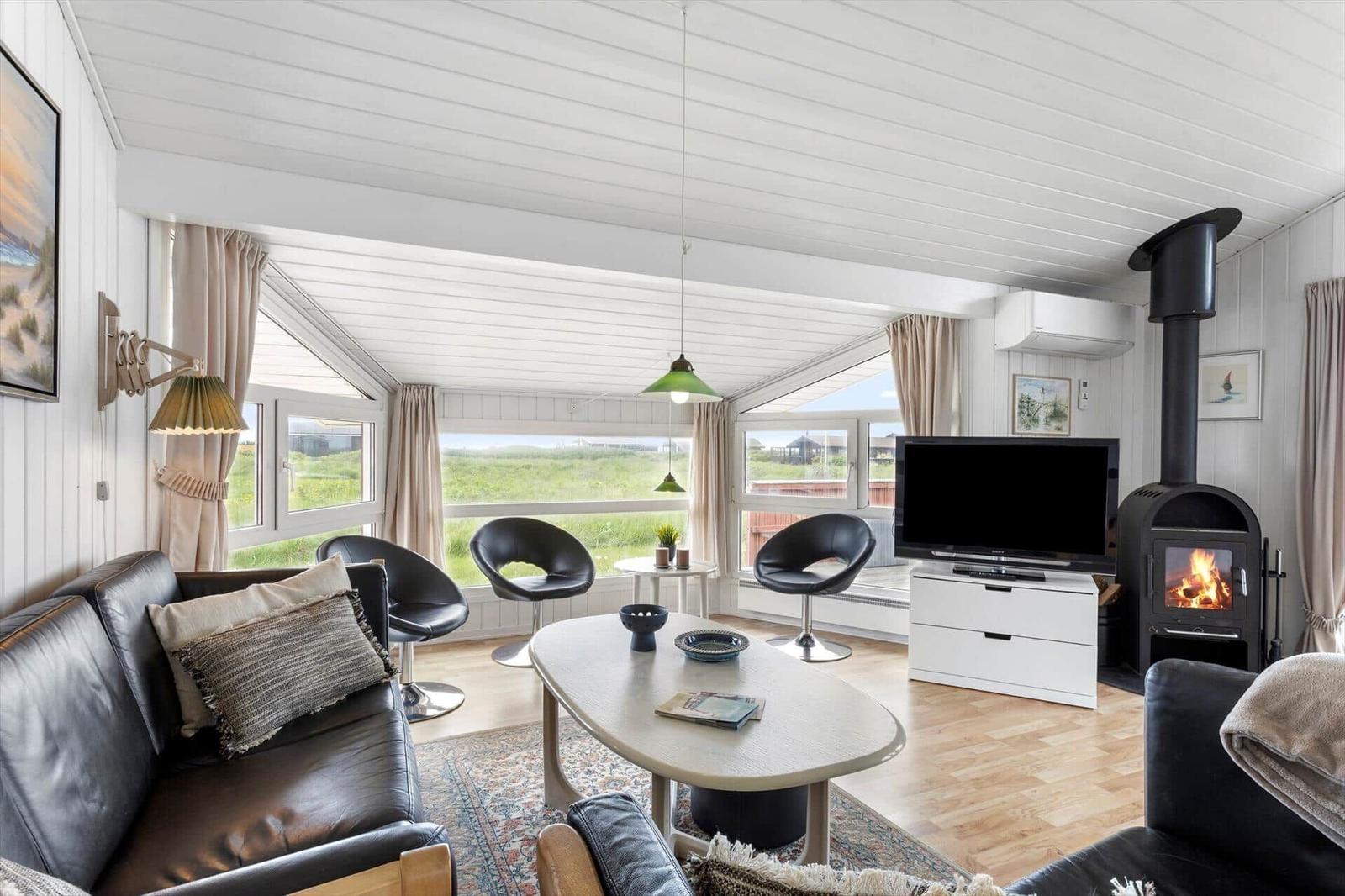 Wohnzimmer mit Sofa, Stühlen, Kamin und TV. Große Fenster mit Blick auf Grünfläche.