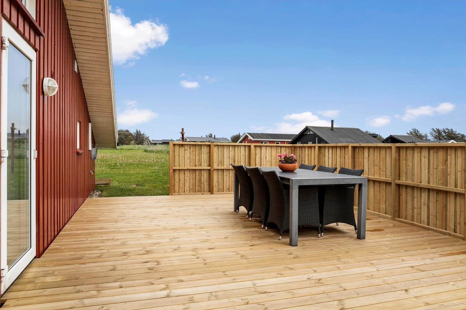 Terrasse mit Holzboden und Esstisch mit sechs Stühlen. Hintergrund: Holzzaun und Grasland.