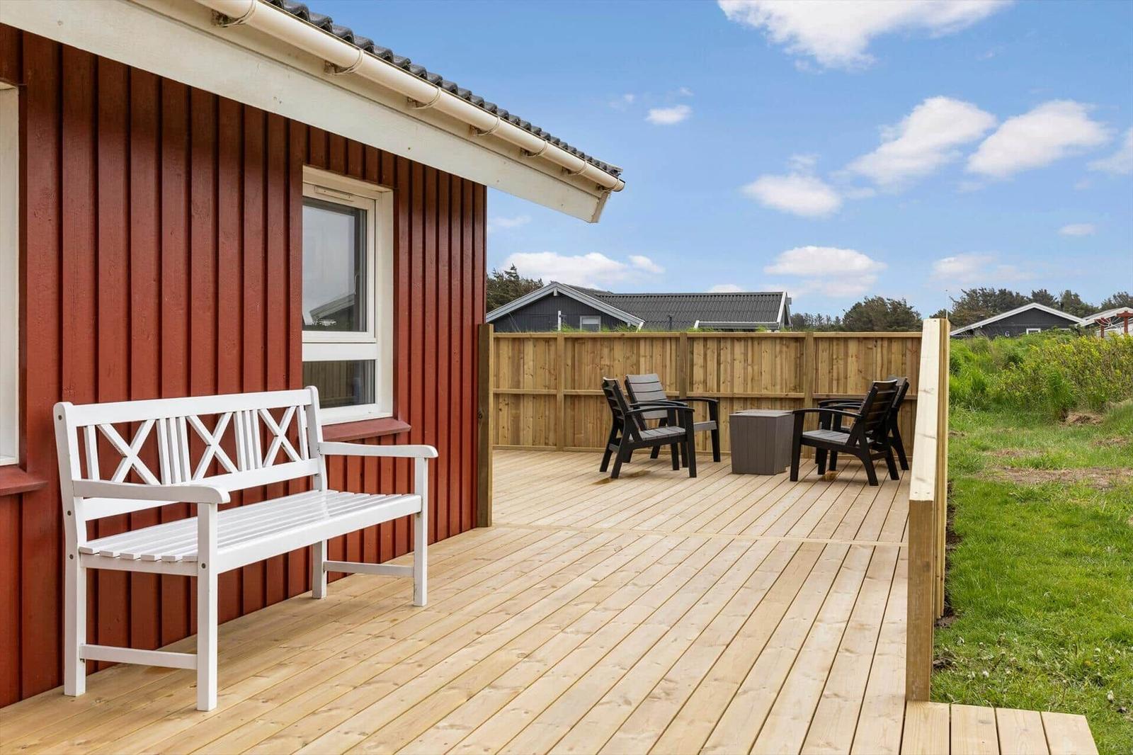 Terrasse mit weißer Bank und Sitzgruppe neben rotem Haus mit Holzgeländer.