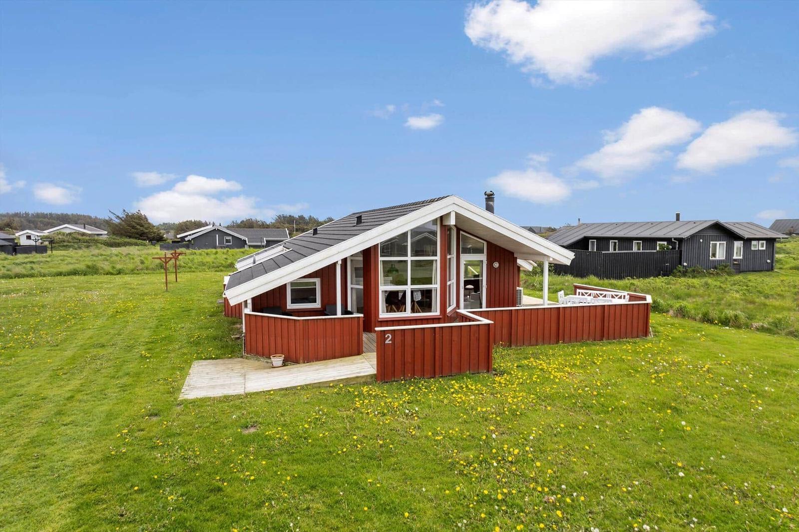 Rotes Ferienhaus mit Terrasse in grünem Grasland unter blauem Himmel.