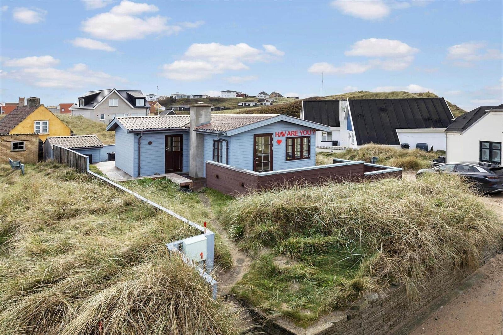 Blue house with red text on the facade, surrounded by dunes and other houses.