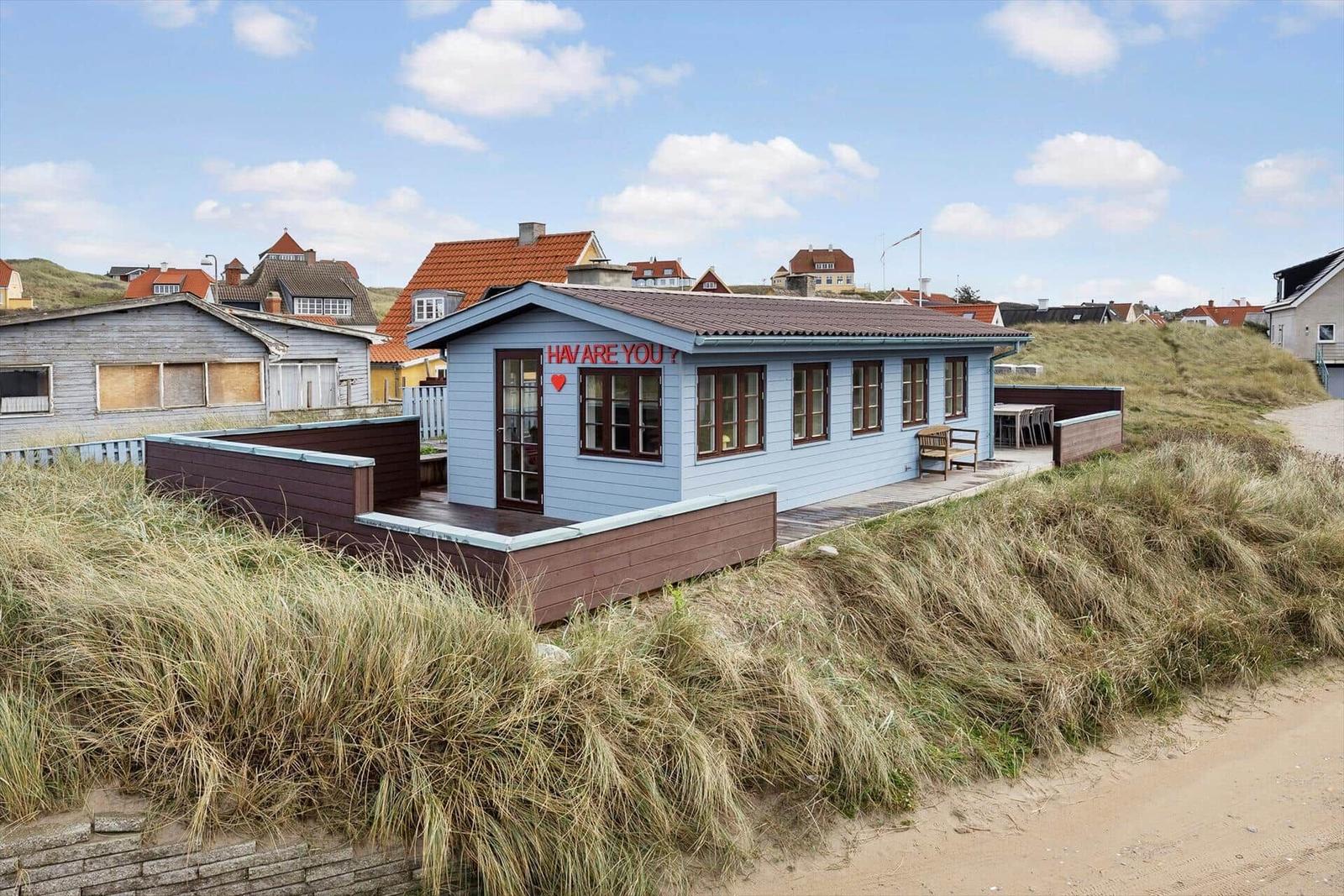 A light blue beach house with terrace and seating area between dunes.