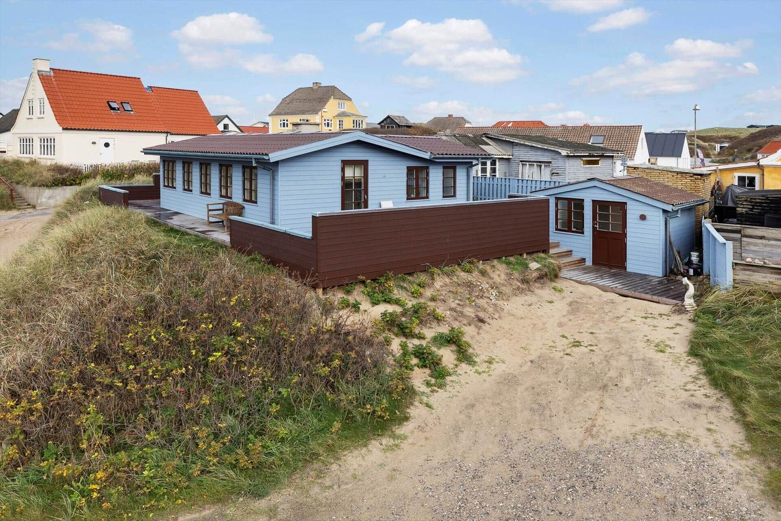 Blue holiday home with brown fence and wooden terrace. Sand path and green areas surround the accommodation.