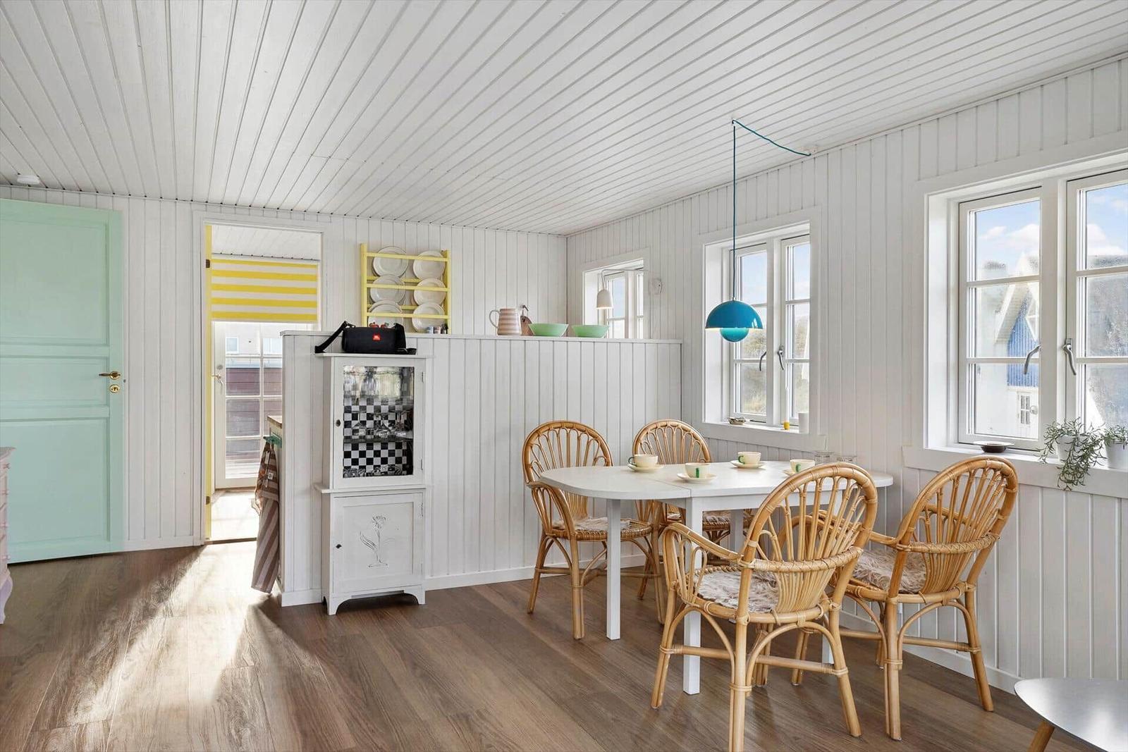 Dining room with round table, rattan chairs, and white wall paneling. Windows offer a view of the outside.