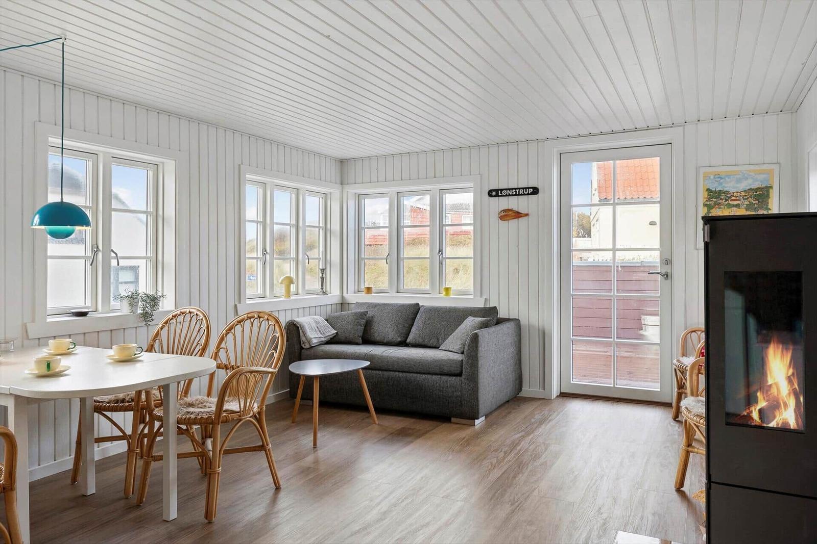 Living and dining area with sofa, table, chairs, and wood stove. Windows and door show outdoor area.