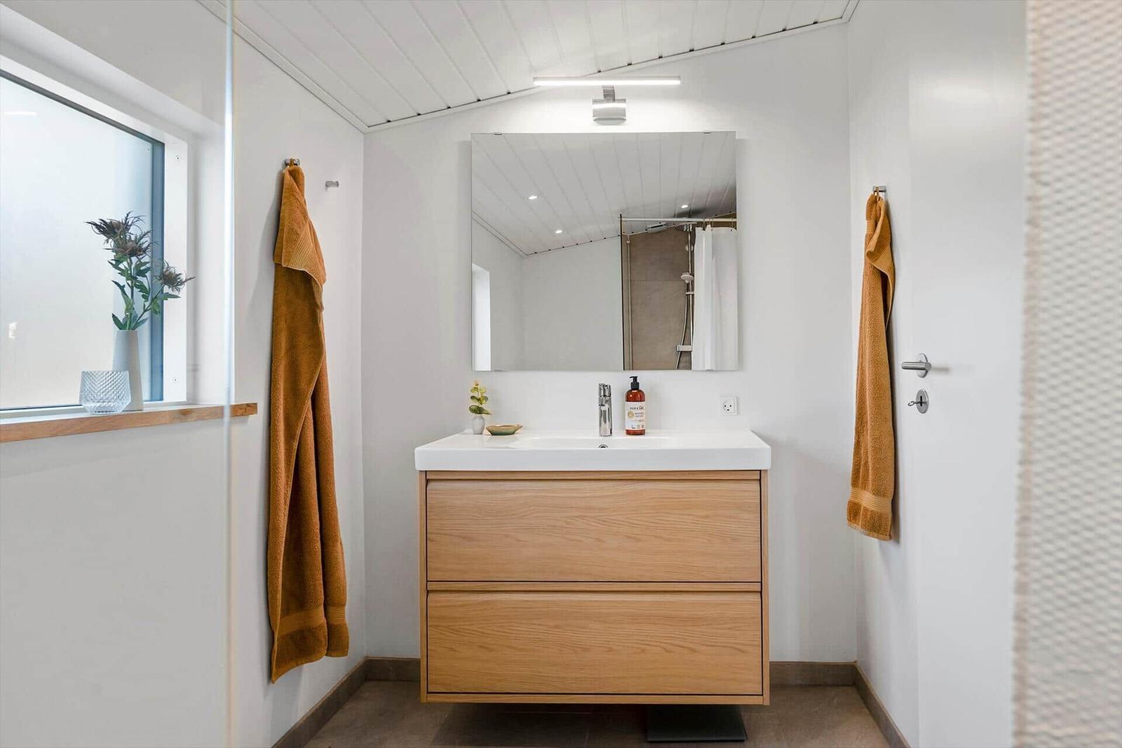 Badezimmer mit Waschbecken, Spiegel und Dusche. Holzschrank, Fenster und Handtücher.