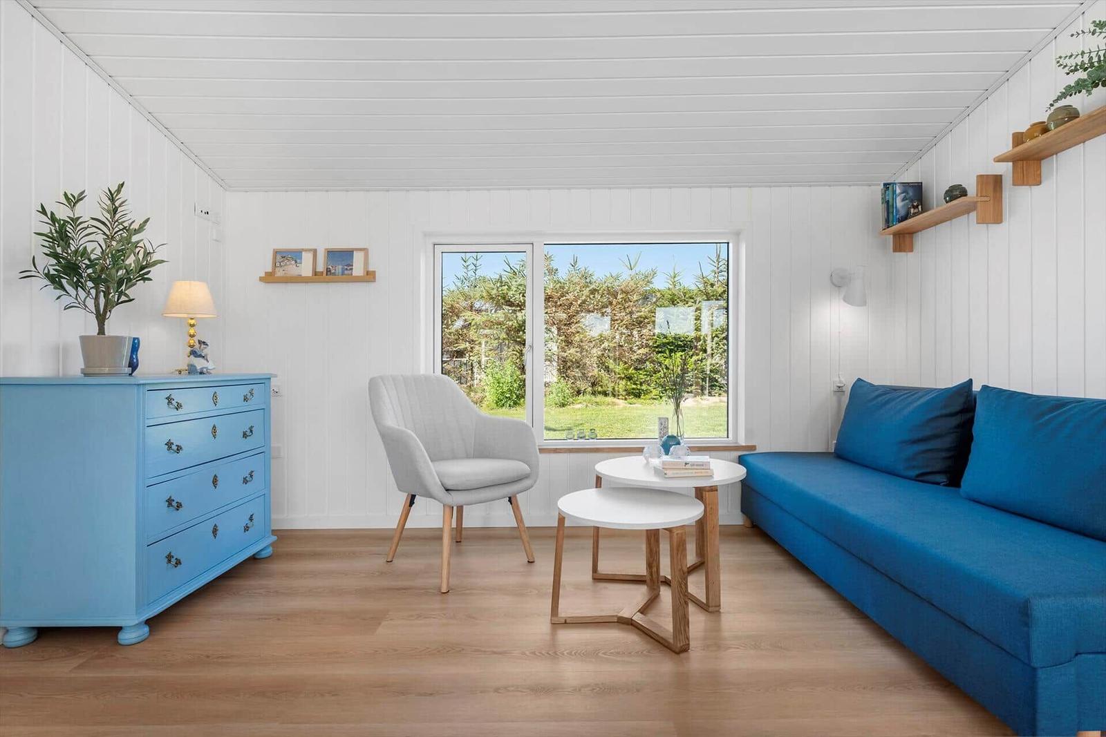 Ein Wohnzimmer mit blauem Sofa, grauer Sitzgelegenheit und Holzboden. Fenster zeigt Garten.