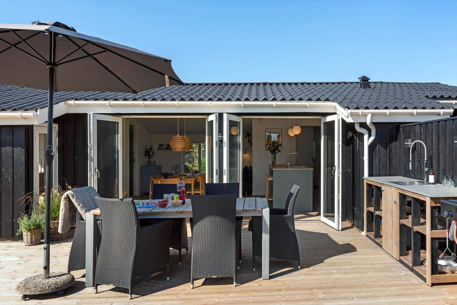 Terrasse mit Tisch, Stühlen und Sonnenschirm. Hintergrund: offene Haustür und Küche.