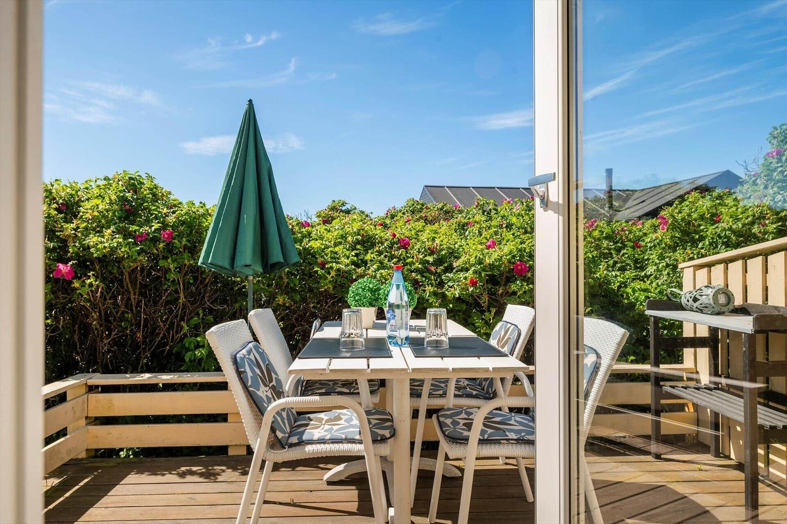 Terrasse mit Tisch, Stühlen und grünem Sonnenschirm. Hintergrund: grüne Hecke mit rosa Blüten.
