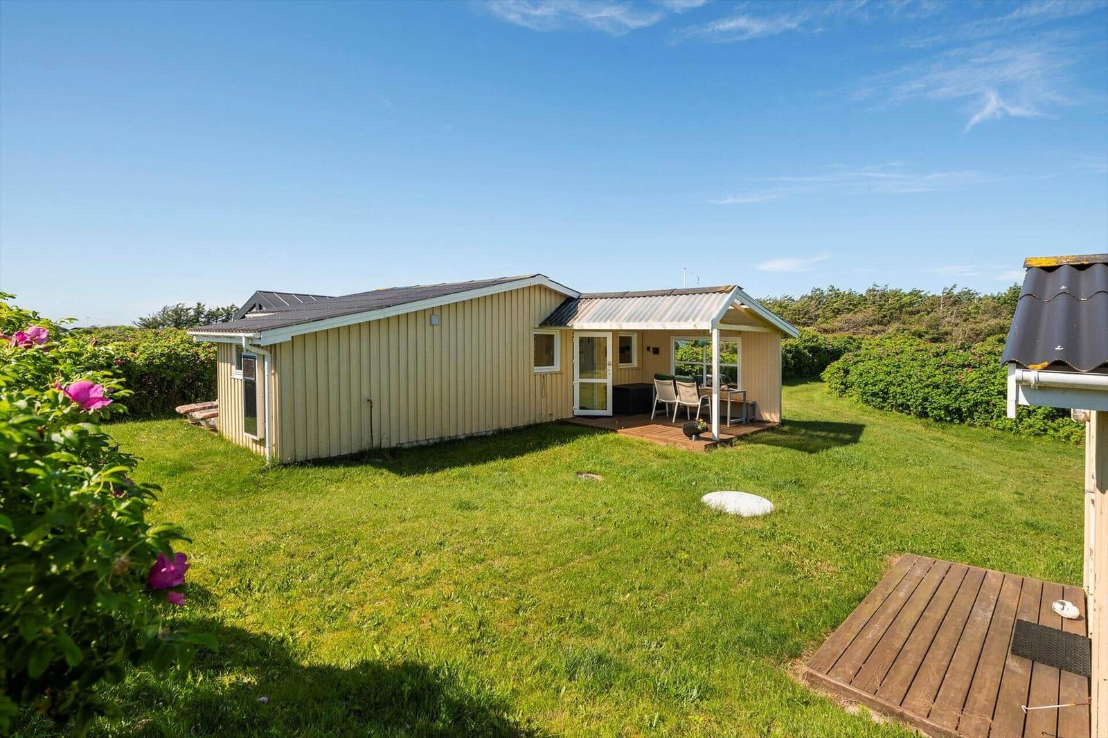 Ein Haus mit Terrasse, Garten und blauem Himmel. Blumen und Gras sind sichtbar.
