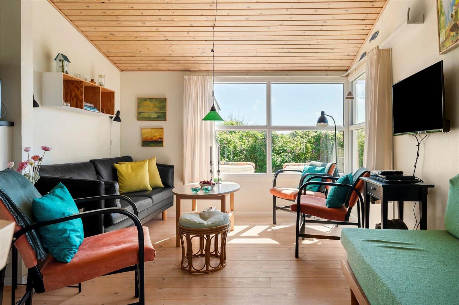 Wohnzimmer mit Holzdecke, Sofa, Sesseln und großem Fenster mit Blick auf Grün.