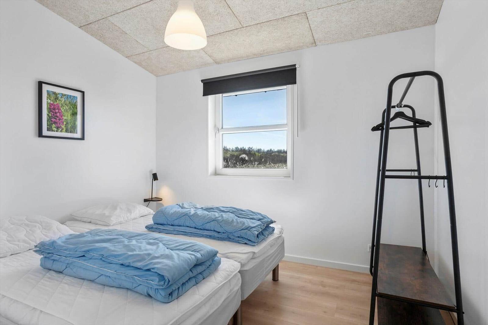 Two beds with blue blankets, window with view of landscape, black coat rack and wall picture.