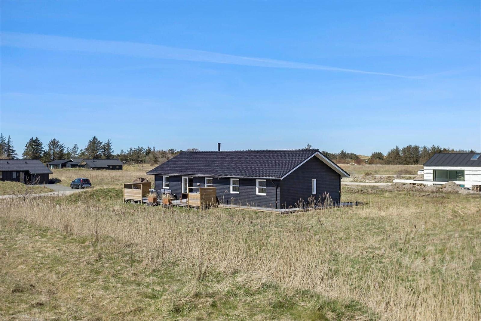 A dark wooden house with a terrace stands in a grassy landscape under a blue sky.