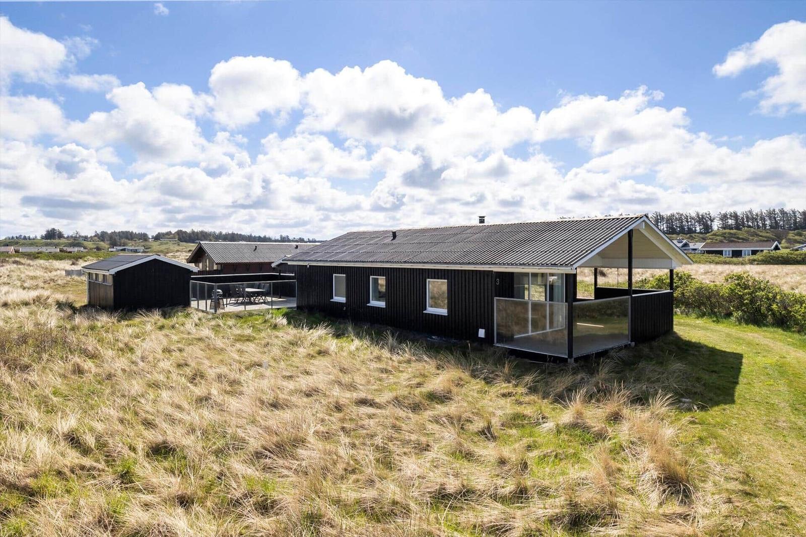 Modern wooden cabin with terrace in a grassy landscape under a blue sky with white clouds.